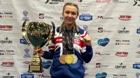 A young woman has her blond hair in two plaits and is wearing a Team GB blue, white and red tracksuit. In one hand she is holding a very large gold cup trophy and her other fist is clenched in victory. She has three large medals around her neck in bronze, silver and gold