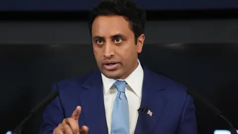 Zia Yusuf is wearing a blue jacket, white shirt and a turquoise-coloured tie. He also has a Union flag pin badge on his lapel.
