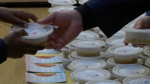 A close up image showing an adult hand, passing a pot of Noah's pudding in a plastic pot into the hands of a child.  There are more pots of pudding on a table in the foreground