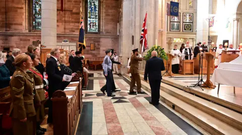 Pacemaker A wide shot of the interior of Belfast Cathedral. In the rows, those in military uniforms watch on as a Union Jack is brought up to the alter of the Cathedral by a uniformed officer. To the side are clergy representatives in white and black robes. The cathedral is made up of white stone walls and marbled forced featuring white, pink and black squares.