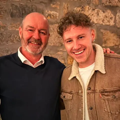 Callum Beattie Steve Clarke, in a navy sweater and white shirt, has his arm around Callum Beattie who is wearing a brown denim jacket and white T-shirt. Both men are smiling in a warm, bright room with a stone wall behind them.