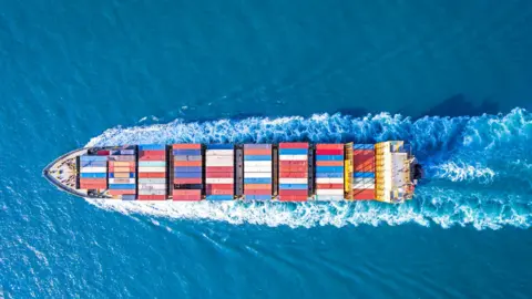 Getty Images A ship carrying shipping containers sailing on the sea. The containers are a range of colours and the sea is a bright blue colour.