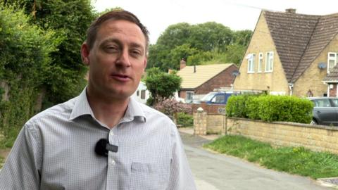 Olly Glover wearing a shirt stood in a residential street with housing behind him