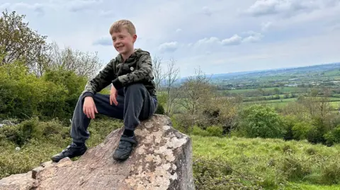 Supplied A young boy in a tracksuit sitting on a rock with a grassy view behind him.