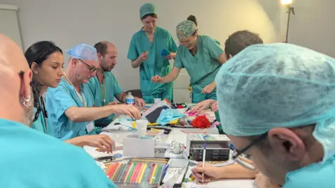 BBC Medics dressed in turquoise scrubs all around a table taking part in an art therapy session, with coloured pens and drawing materials on the table
