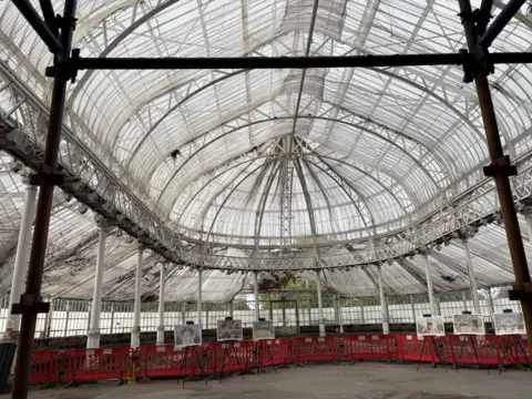 An image of a huge class house in daylight, clearly in the midst of construction work - with red barriers separating the floor area, and netting stretched across the ceiling under the glass panes.