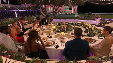 REX/Shutterstock A group shot of Love Island contestants sitting around a round dinner table, with food on plates, flowers and a white tablecloth.