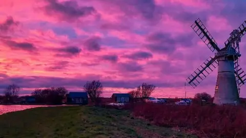 A windmill on the banks of the River Thurne with riverside properties in the background. The sky is a mixture of pink and purple