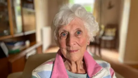 An elderly woman with grey hair and blue eyes in a blue, pink and white top. 