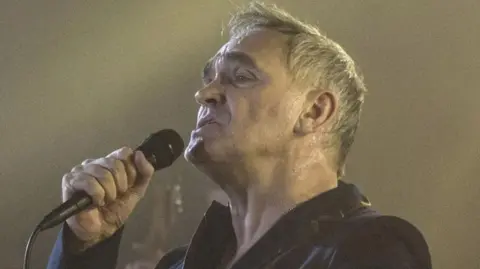 BBC/JAMIE SIMONDS Morrisey holds a  microphone on stage wearing a black jacket