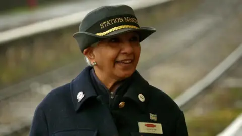 Chandra is wearing a blue wool coat with a white name badge and a grey hat with 'Station Master' in gold lettering