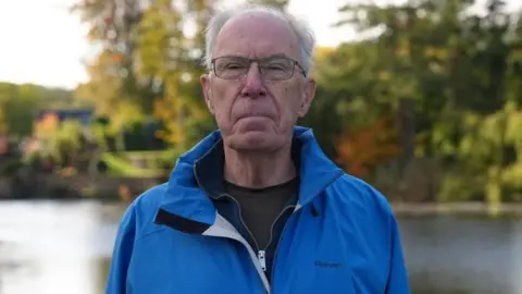 A man wearing a blue coat looks into the camera with a neutral expression on his face. He is standing next to a river.