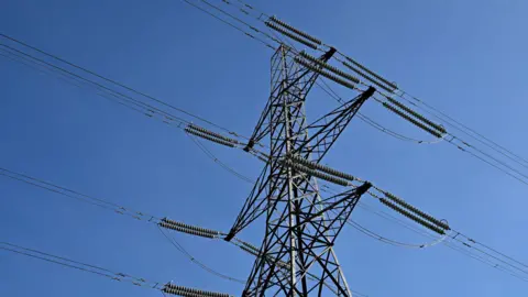 An electricity tower, or pylon, pictured looking up it from the ground and against a clear blue sky. The tower is a lattice-work of metal with "arms" sticking from the side attached to cables.