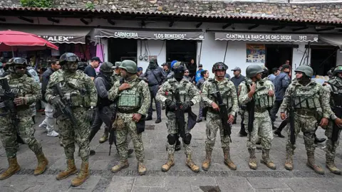 Mexican security forces seen carrying out an anti-cartel raid last month. They wear military apparel and have their faces covered. There's a butcher's shop in the background