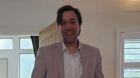 Family A man with brown wavy hair, a pale pink suit jacket and a white shirt smiles at the camera