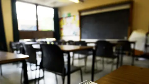 A generic shot of a classroom with rows of chairs tucked into individual desks. At the front of the room is a blackboard. 