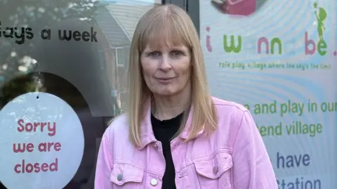 A woman with shoulder length blonde hair and a blonde fringe is standing next to a large glass window. She is wearing a pink jacket and a black top. There is a sign saying "Sorry we are closed" next to her on the window.