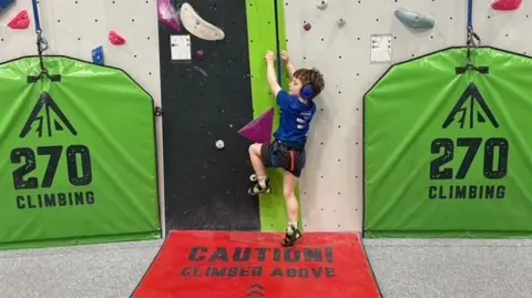 A young boy in a blue top, black shorts and black shoes. He is attached to a rope and is about to start climbing up a climbing wall. There are two green signs either side which say '270 climbing'. He is stood on a red sign saying 'caution climber above'. 