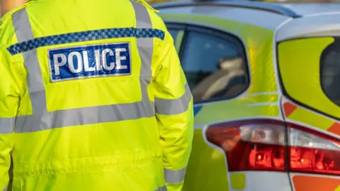The back of a police officer in a fluorescent yellow jacket, with the word POLICE printed on the jacket. In front of the officer, is a marked police car, which is yellow, blue and orange.