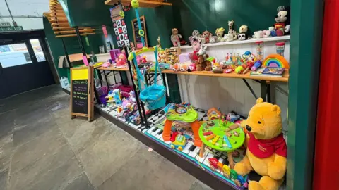 An array of children's toys from a Winnie the Pooh cuddly toy to a toy phone are displayed on three shelves. The wall behind the display is teal and has vertical white boards along the bottom half.