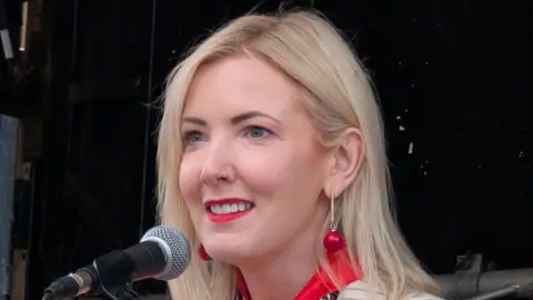 Sharon has long blonde hair, red earrings and a red top with red lipstick.