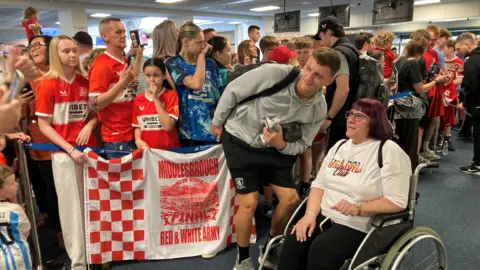 A crowd of people, three or four deep, stand behind a barrier in the airport to greet Middlesbrough players. Dael Fry, who is wearing a grey jumper and black short, is slightly bent over to pose for a photo with a person in a wheelchair who has mid-length dark purple hair.
