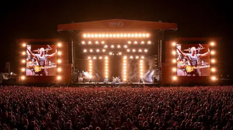 The BBC Radio 2 in the Park stage, as well as a large crowd standing in front of it, is illuminated by bright lights. 