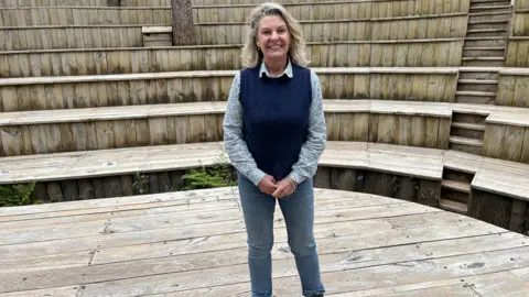 Darren Rozier/BBC A woman stands at the bottom of an outdoor theatre. She has shoulder length blonde and grey hair and she wears a blue floral shirt with the navy sleeveless jumper over the top along with jeans. She rests her hands in front of her body as she smiles.