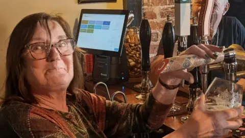 Sally Ward standing behind the bar of a pub pulling a pint. She is looking directly at the camera and is wearing a green, orange and brown patterned long sleeved T-shirt. She has a pair of glasses on her eyes.