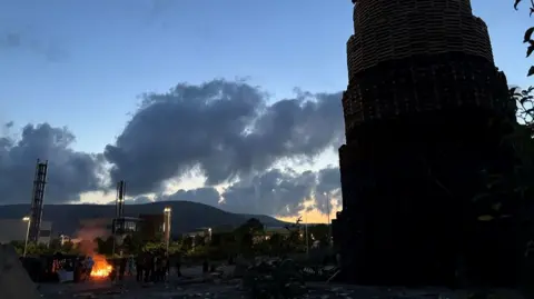 The south Belfast bonfire at dusk, with small crowds of people around it and a small fire has been lit beside the bonfire. 