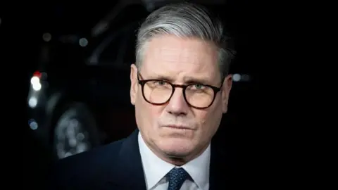 A close up of Keir Starmer's face. He is wearing a dark suit and glasses and has a serious expression on his face 