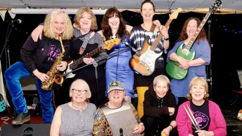 The HorMones The nine women pose with their instruments on a stage. Instruments include guitars, bass, saxophone, drums and a washboard.