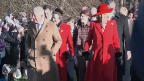 BBC King Charles dressed in a beige long coat walks along side Queen Camilla dressed in red