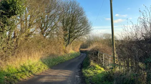 A photo of Batley Lane, in Pleasley. A thin single-track road can be seen on the right. On the left are two triangle road signs of a person riding a horse, and another of a bicycle