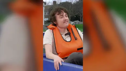 Sam Campbell has brown hair and is wearing an orange life-jacket