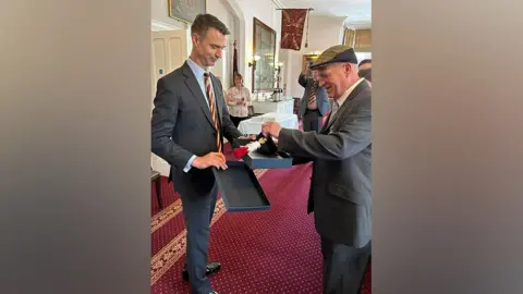 The Royal Regiment of Fusiliers Major General Jon Swift, dressed in a dark grey suit, presents Fusilier Steve Close, also wearing a grey suit, with a presentation box containing his beret and hackle, made up of red and white feathers. Several people are smiling in the background