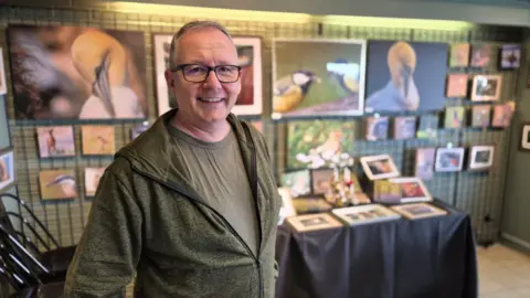 Stuart Woodward/BBC Alan Bruce stands in front of a selection of his wildlife photographs which are on display at Fingringhoe Wick Nature Discovery Park. He is wearing a green hoodie over a green T-shirt. He has dark-rimmed glasses and is smiling at the camera