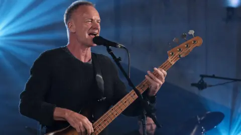 BBC/Michael Leckie A wide shot of Sting playing a bass guitar. He has short grey hair and is wearing a black jumper.