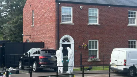 Tow men in police forensic suits stand outside a red brick house. A black car and a white van are parked outside.