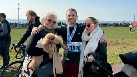 Brighton Half Marathon A man stood with two women and a dog. The man is wearing a medal. 