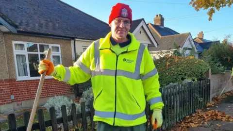Greener Ealing A man wearing a fluorescent jacket and red hat and a broom handle stands in front of a bungalow.