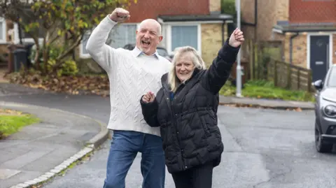 Man in white jumper and woman in dark coat both with hands in the air in celebration stading in a cul de sac with houses behind them
