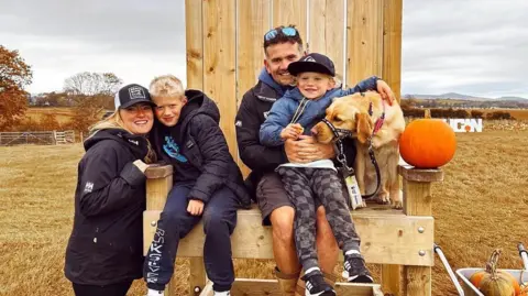 Andy Johnson Photograph of Andy and Danni Johnson with their children, Finley and Oakley and their guide dog, Pearlie. Andy, Finley, Pearlie and Oaklen sit on a large wooden chair and Danni stands next to the chair smiling. 
