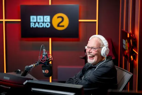 Sarah Louise Bennett/BBC Bob Harris sits in the Radio 2 studio, with its logo on the microphone and walls, wearing white headphones and laughing into the microphone. The walls are red with orange lighting.