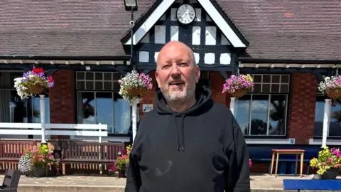Stockport Cricket Club Herman stands outsdie the club house, smiling. It is sunny. 