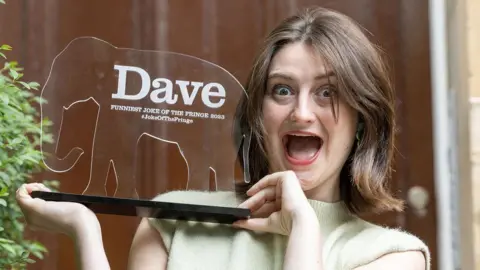 PINPEP/Robert Perry Female comedian Lorna Rose Treen holds trophy with a wide grin expression on her face