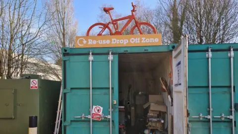 FCC Environment Front of an open old shipping container, painted green. Various items inside and boxes. On the top, an old orange painted bike and a sign saying 're-use drop off zone'.