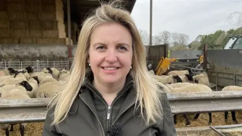 Isobel smiles for the camera, her sheep in a pen behind her. She has long blonde hair and wears a browny green jacket and fleece.