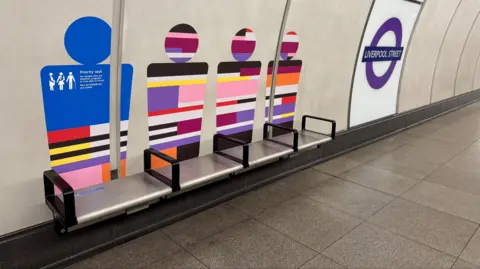 Colourful outline of people on wall of Liverpool Street Elizabeth line platform above silver seats with black armrests