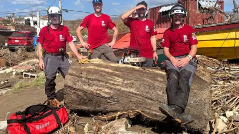 supplied Four men in red t-shirts sit and stand on a large tree trunk in the sunshine. A chainsaw is resting on the trunk.